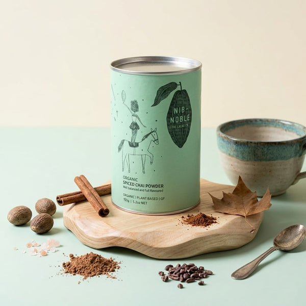 Canister of Nib & Noble Spiced Chai Powder on a wooden board with spices and a cup.