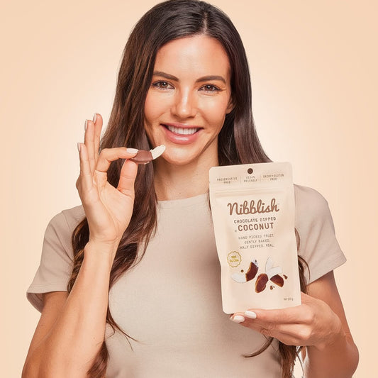 A smiling woman holds a small piece of chocolate-coated coconut between her fingers while holding a stand-up pouch labeled 'Nibblish Chocolate Coffee Coconut' against a warm beige studio background.