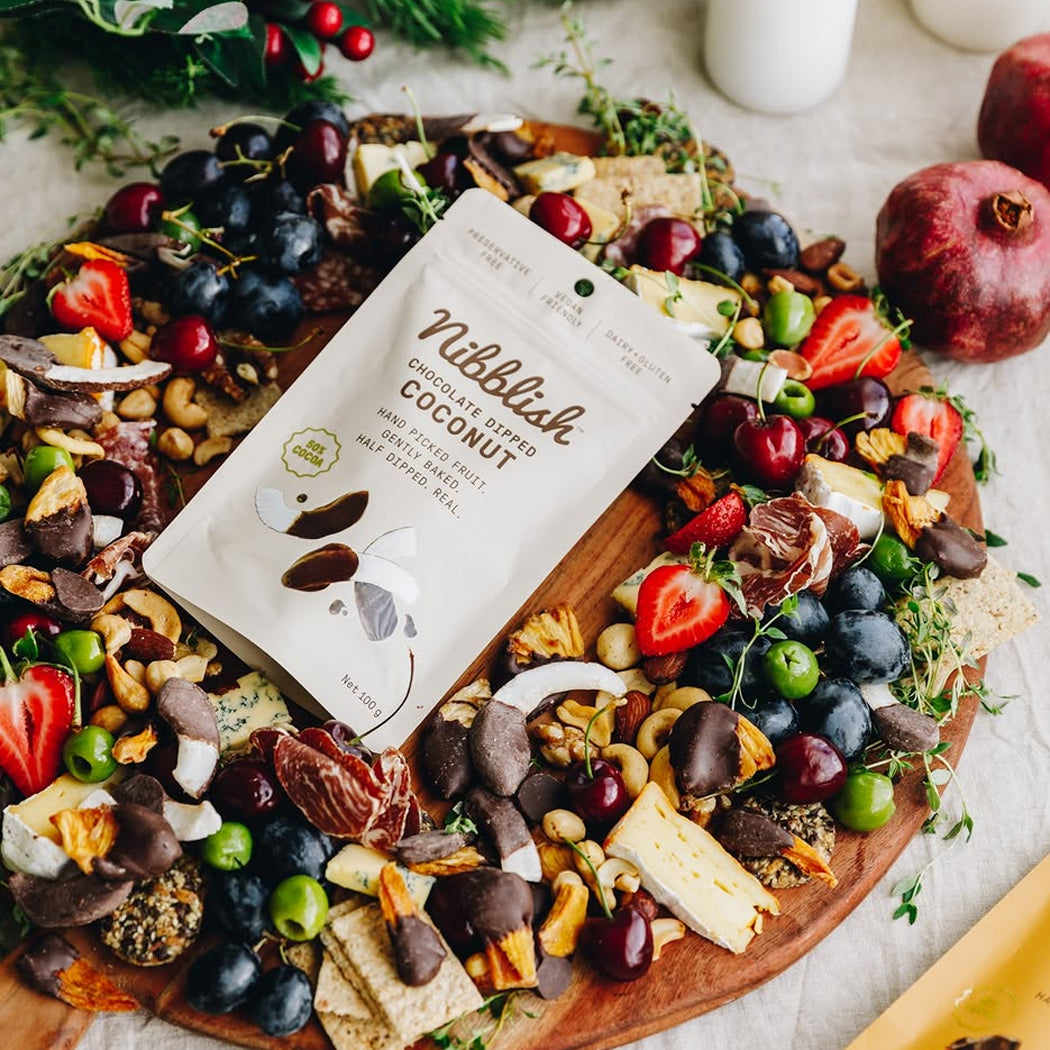 A stand-up pouch of Wilblisk Coconut Flavored Snacks is placed on a large wooden charcuterie board filled with nuts, dried fruit, crackers, blueberries, strawberries, and cheese, with pomegranate and greenery around it.