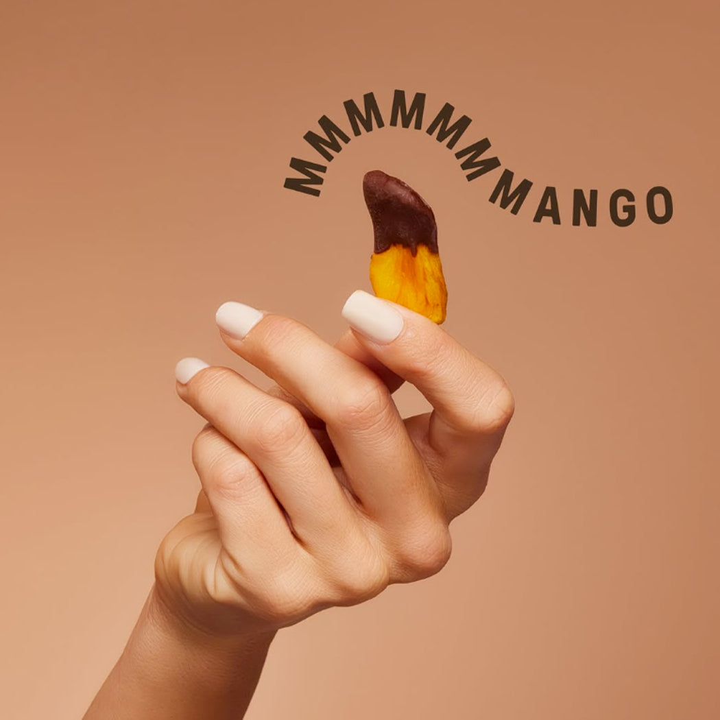 A hand with white manicured nails holds a small piece of mango that is dipped half in chocolate. The background is a solid warm peach color with the text 'MMMMMMANGO' written in a curved black font above the hand.
