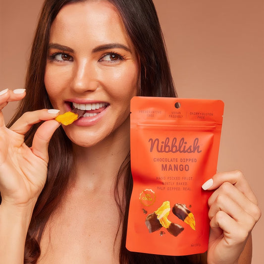 A smiling woman holds a bright orange Nibblish Chocolate Dipped Mango pouch while eating a piece of the yellow mango dip. The background is a solid warm beige studio backdrop.