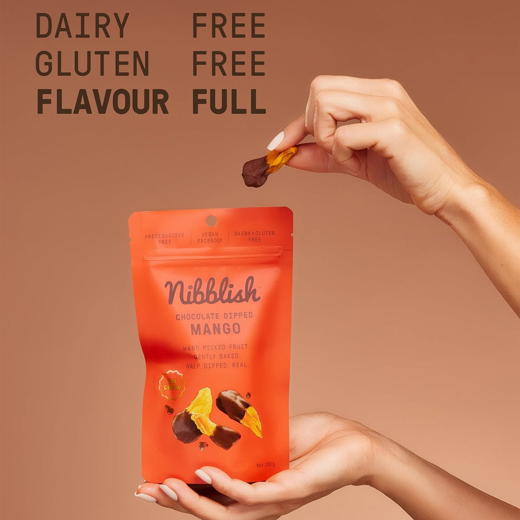 A person holds an orange stand-up pouch of Nibblish Chocolate Dipped Mango sticks while another hand lifts a chocolate-dipped mango stick above the package. The background is a solid warm tan, with text reading 'DAIRY FREE GLUTEN FREE FLAVOUR FULL' at the top.