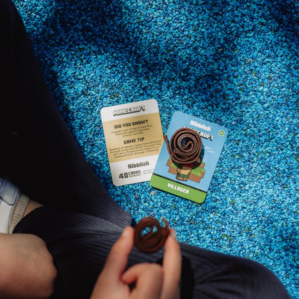 A person holds a small brown coiled item in their hand while sitting on a bright blue carpeted floor. Two cards are placed nearby on the floor: one white card with a gold-toned panel reading 'PUBG: BLOODSTRAIN' and 'GRACE TIP', and one blue card with a cartoon illustration and the word 'VILLAGE'.
