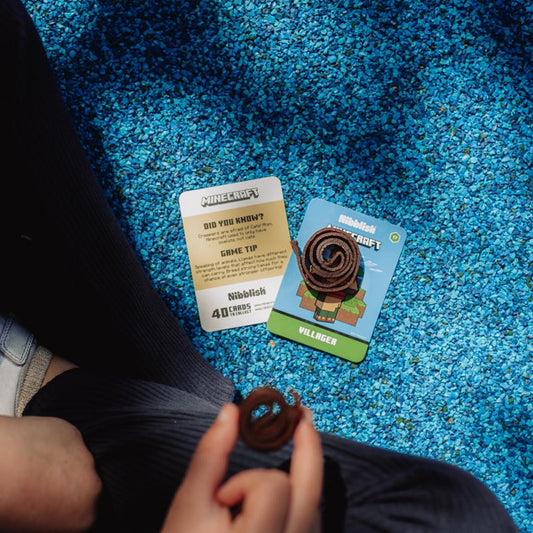 A person holds a small brown coiled item in their hand while sitting on a bright blue carpeted floor. Two cards are placed nearby on the floor: one white card with a gold-toned panel reading 'PUBG: BLOODSTRAIN' and 'GRACE TIP', and one blue card with a cartoon illustration and the word 'VILLAGE'.