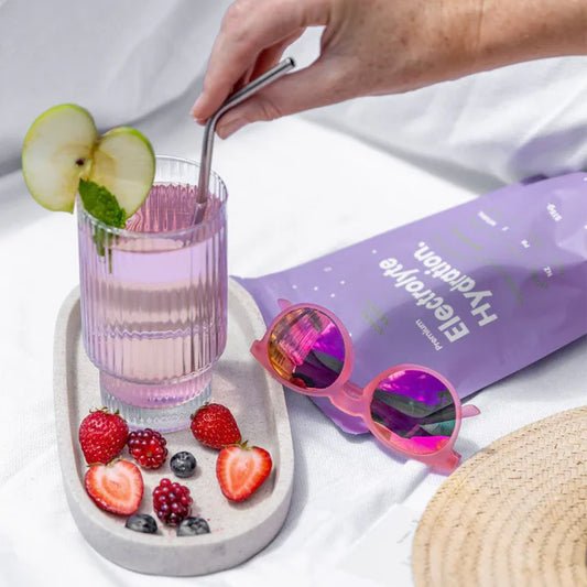 Person stirring a pink drink in a glass with fruit slices, next to a purple energy drink package and sunglasses.