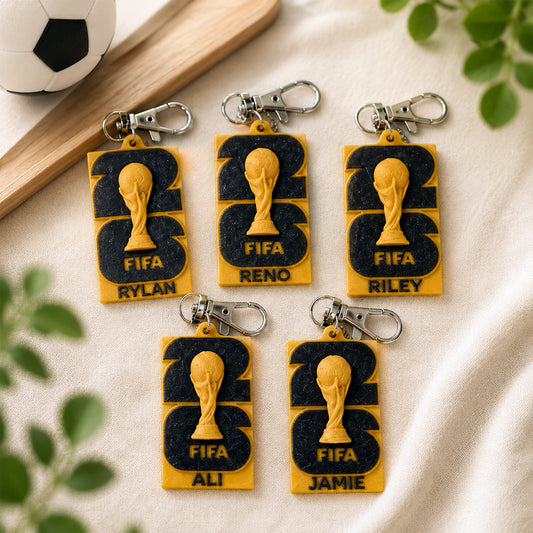 Five football-themed keychains arranged on a light fabric surface, each with a gold trophy graphic, dark navy lettering, and a silver key ring. Decorative leaves, a wooden board, and part of a soccer ball appear around the edges.