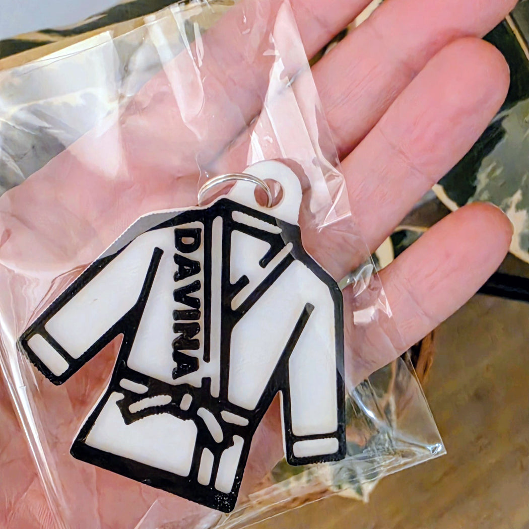 A white and black EVA keychain shaped like a martial arts gi jacket with the word 'DAYNA' printed vertically, held in a hand inside a clear plastic bag.