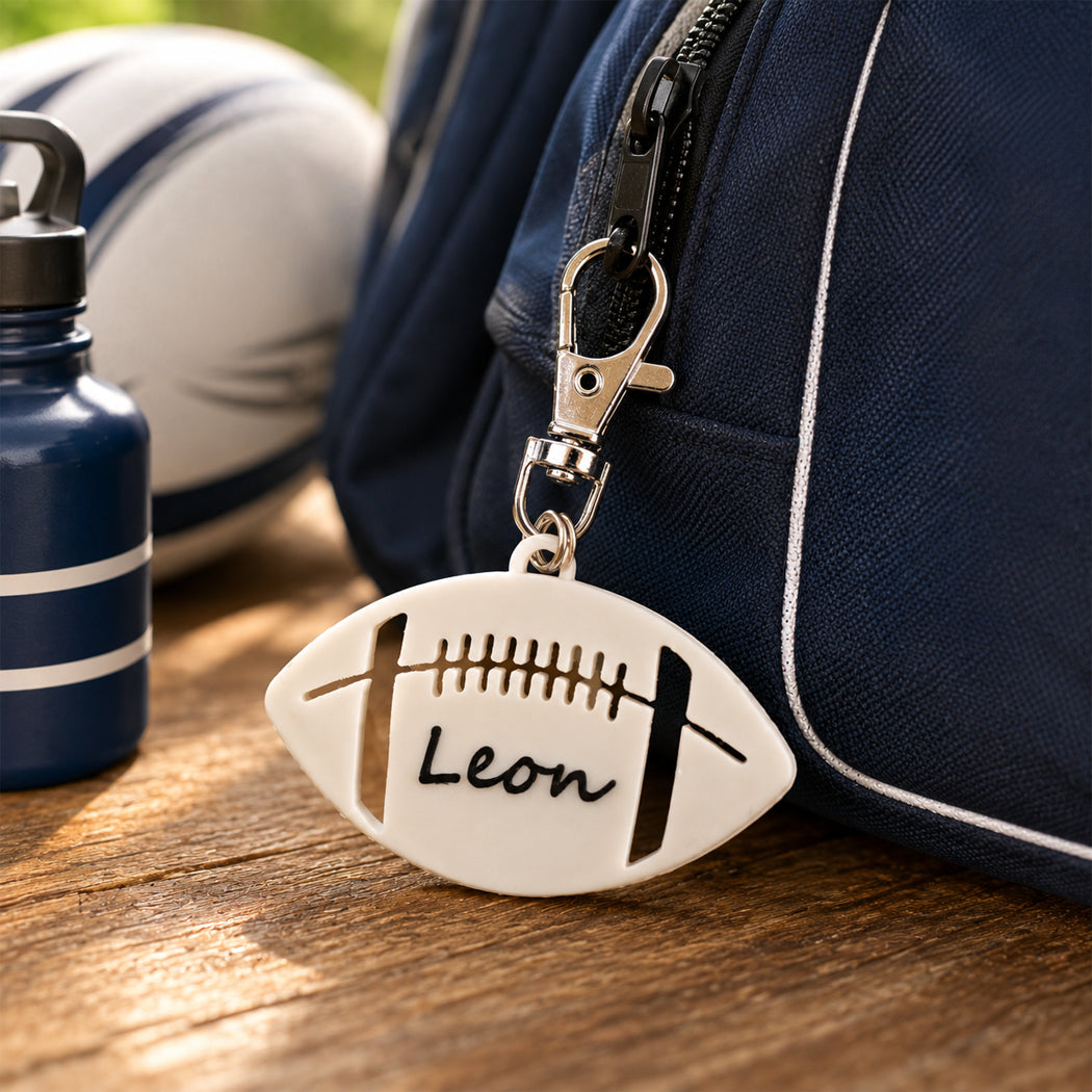 A football-shaped keychain charm with the name 'Leon' engraved, attached to a navy blue bag and resting on a wooden surface next to a water bottle.