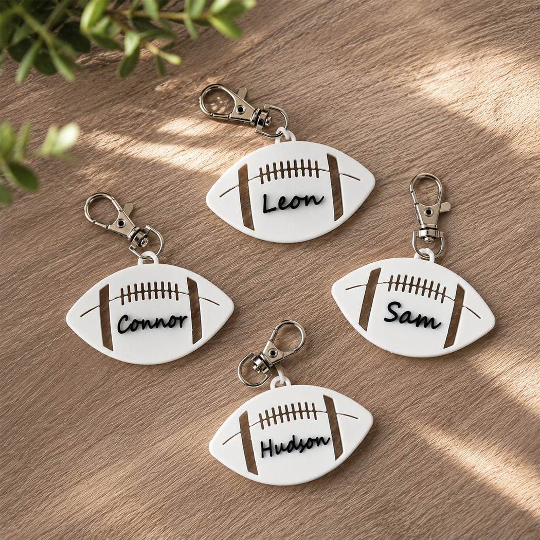 Four personalized football-shaped keychains with silver keyrings are arranged on a wooden surface with soft leaf shadows. The keychains are white with brown laces and names engraved: Leon, Connor, Sam, and Hudson.