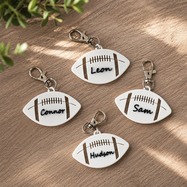 Four personalized football-shaped keychains with silver keyrings are arranged on a wooden surface with soft leaf shadows. The keychains are white with brown laces and names engraved: Leon, Connor, Sam, and Hudson.
