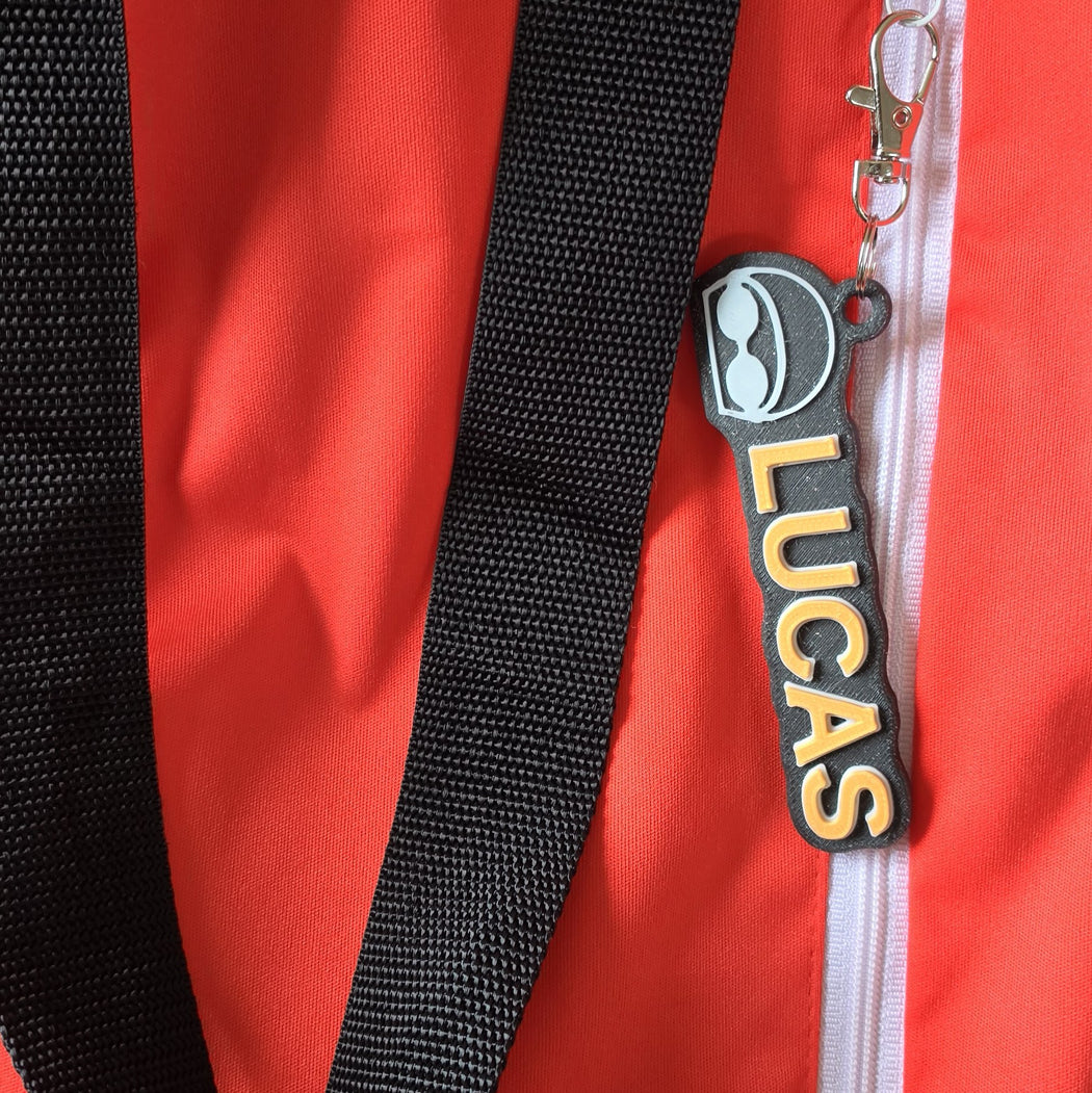 Close-up of a red zippered bag with a black webbed strap and a black zipper pull featuring the name LUCAS in yellow letters.