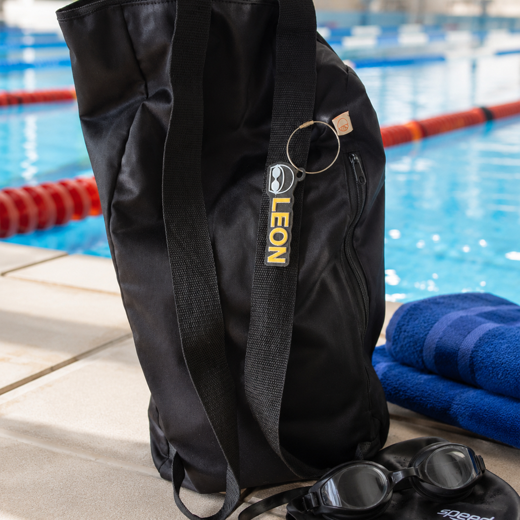 A black athletic tote bag with a vertical 'LEON' logo tag, standing on a pool deck next to blue towels and swim goggles, with a swimming pool in the background.
