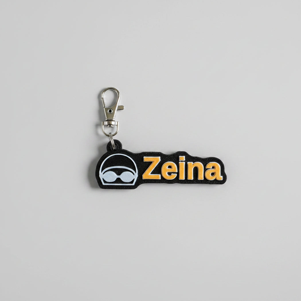 A black silicone keychain charm featuring the name 'Zeina' in gold lettering with a white outline, alongside a black and white swim goggles icon. The charm is attached to a silver metal lobster clasp and photographed on a neutral light gray background.