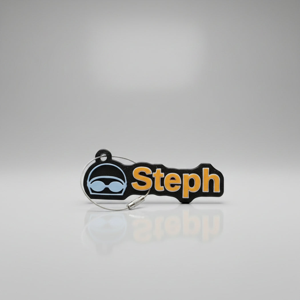 A custom black rubber name tag with the name 'Steph' in orange lettering and a light blue goggles icon, shown on a reflective gray background.