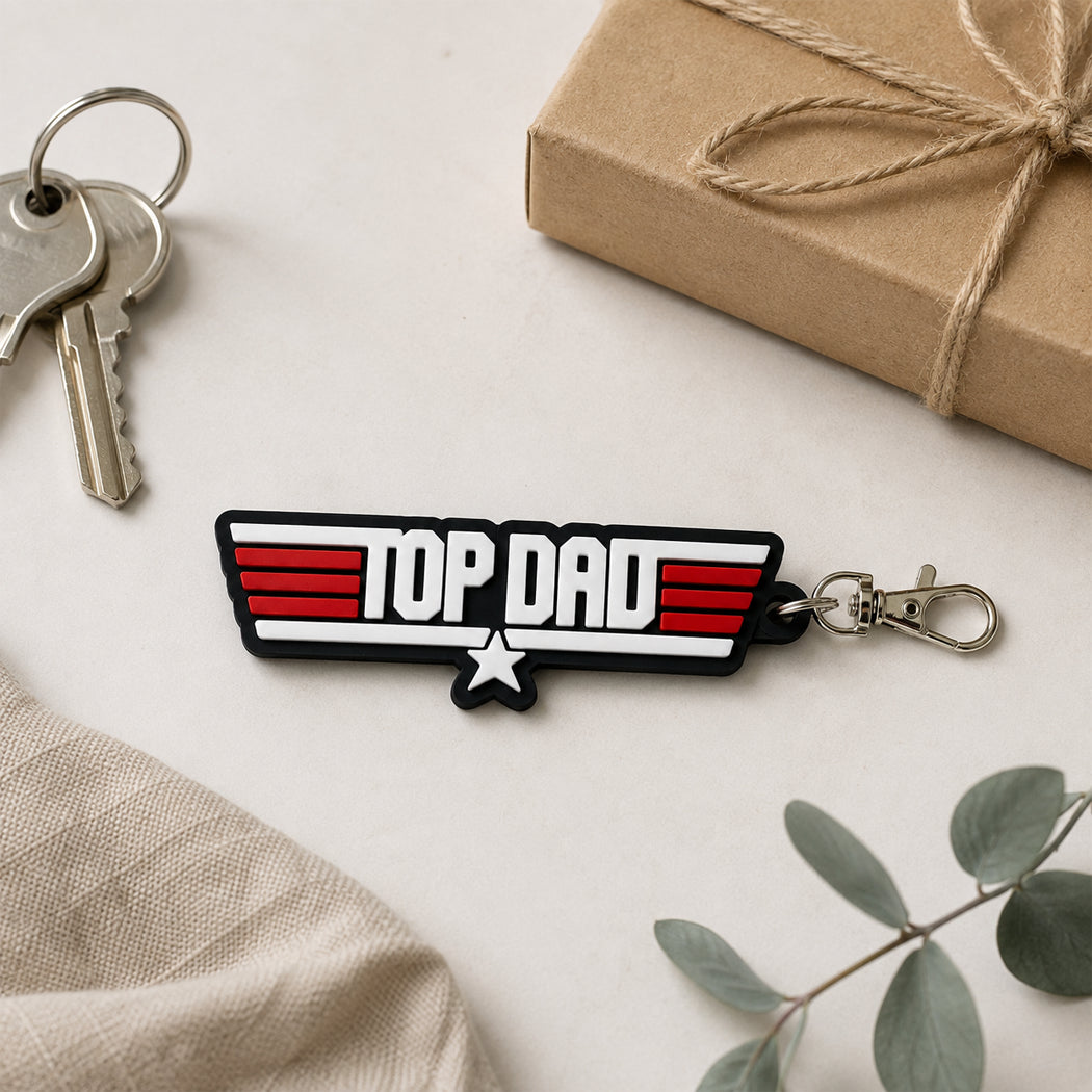 A 'Top Dad' themed keychain with a retro military-style insignia design in red, white, and black, featuring a silver clasp. The keychain is styled in a flat-lay arrangement with keys, a brown kraft gift box, and a beige linen cloth on a light cream background.