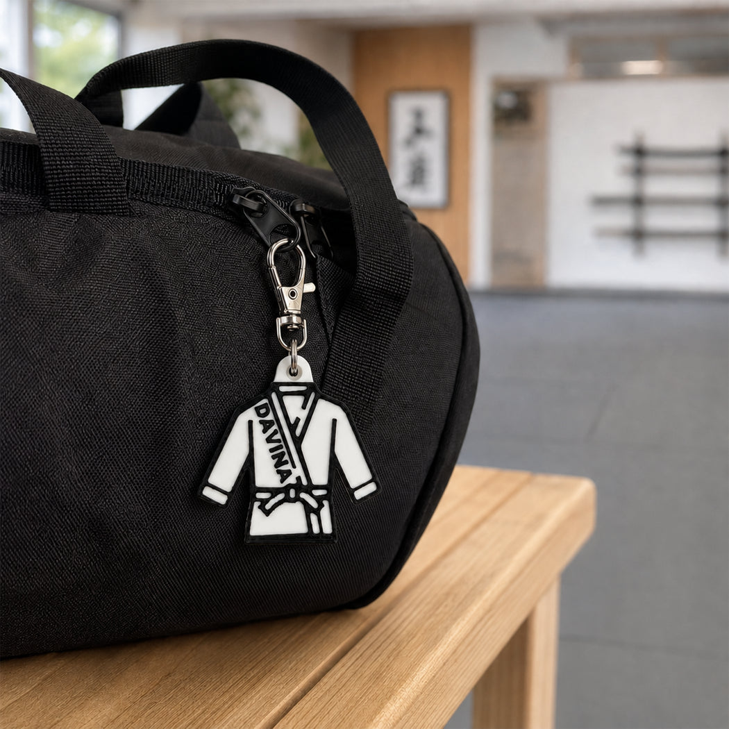 A black fabric duffel bag sits on a light wood table in a blurred indoor space. Attached to the zipper is a white and black martial arts uniform keychain charm with the text 'DIVANA' on it.