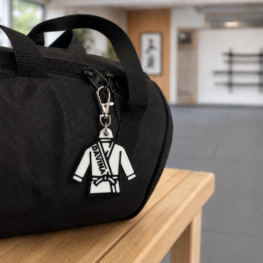 A black fabric duffel bag sits on a light wood table in a blurred indoor space. Attached to the zipper is a white and black martial arts uniform keychain charm with the text 'DIVANA' on it.