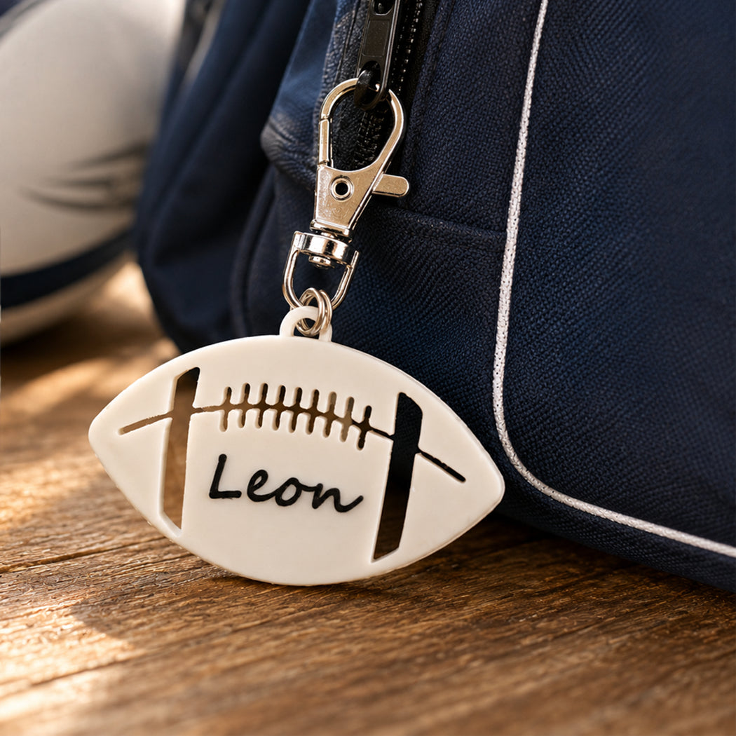A custom white football-shaped keychain with black lacing details and the name 'Leon' written in black script, attached to a silver clasp and shown next to a navy blue bag on a wooden surface.