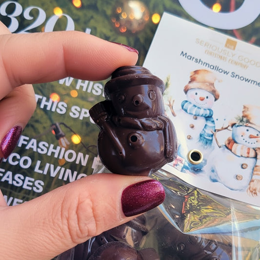 Chocolate snowman-shaped item held by a hand with purple nail polish, against a blurred magazine background.