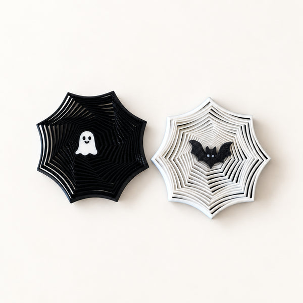 Two Halloween-themed fidgets on a light cream background. The left decoration is black with a white ghost in the center, and the right decoration is white with a black bat in the center.
