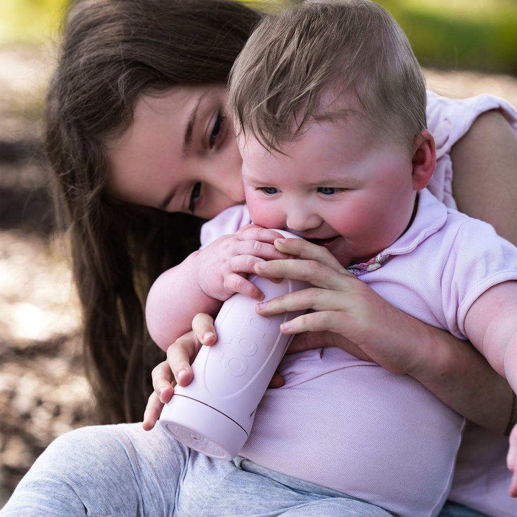 Woman holding a baby who is drinking from a pink sippy cup outdoors.