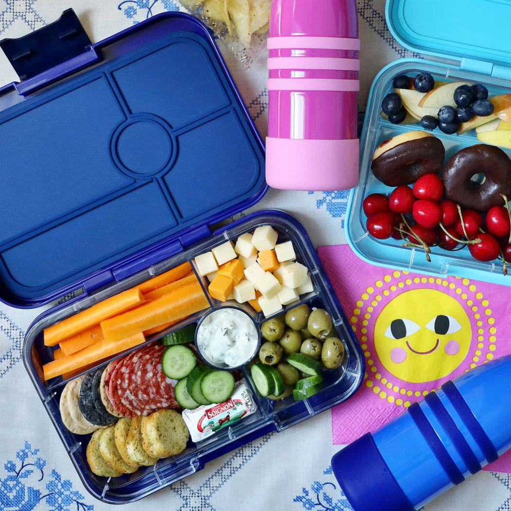 Open blue bento box with snacks and a pink water bottle on a patterned tablecloth.