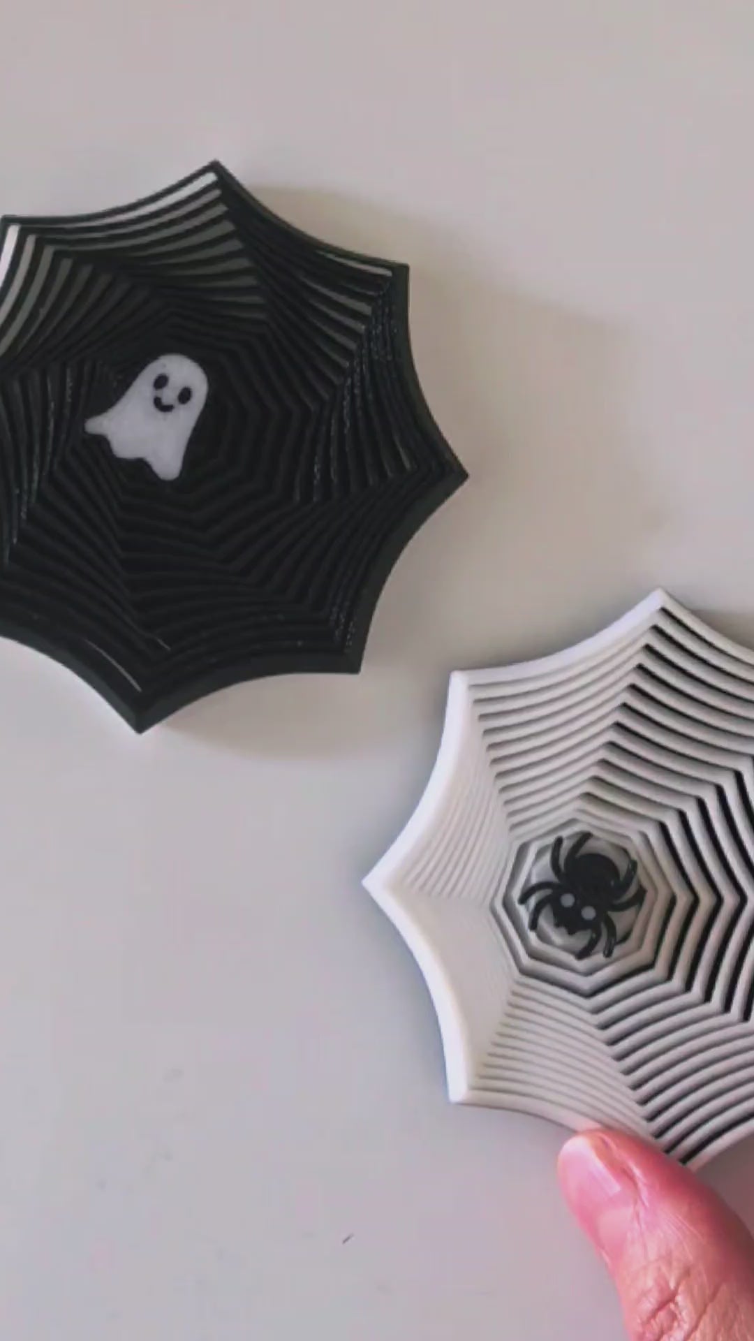 Two Halloween-themed fidgets on a light cream background. The left decoration is black with a white ghost in the center, and the right decoration is white with a black bat in the center.